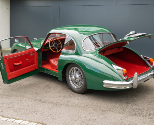 Ein grüner Jaguar XK150 mit rotem Interieur steht mit offenen Türen und Kofferraum vor einer Garage.