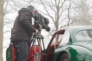 Ein Kameramann von hinten. Er steht neben einem Jaguar XK 150 mit offener Türe, und filmt durch diese das Interieur des Fahrzeugs. Im Hintergrund leicht vom Nebel verhüllte spätwinterliche Bäume.