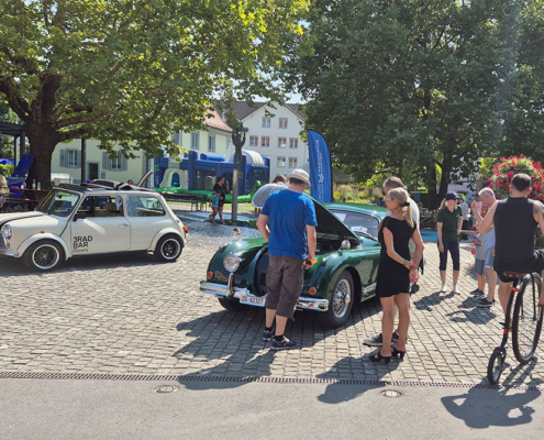 Leute stehen um einen grünen Jaguar XK150 mit offener Fronthaube. Jemand fährt auf einem alten Hochrad vorbei.