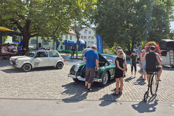 Leute stehen um einen grünen Jaguar XK150 mit offener Fronthaube. Jemand fährt auf einem alten Hochrad vorbei.