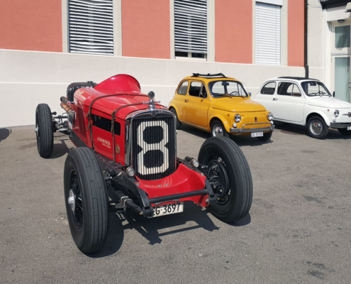 Links ein roter Nash Twin Ignition Eight in offener Bauweise. Sein Kühlergrill wird von einer 8 geziert. Rechts davon zwei Fiat Nuova 500, einer in gelb, einer in weiss.