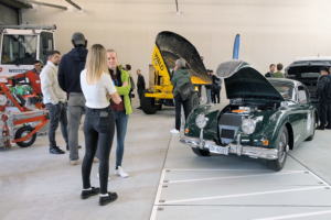 Verschiedene Fahrzeuge, unter anderem ein Muldenfahrzeug, ein Mäher sowie ein Jaguar XK 150 stehen in einem Hangar. Diverse Menschen betrachten und besprechen die Fahrzeuge