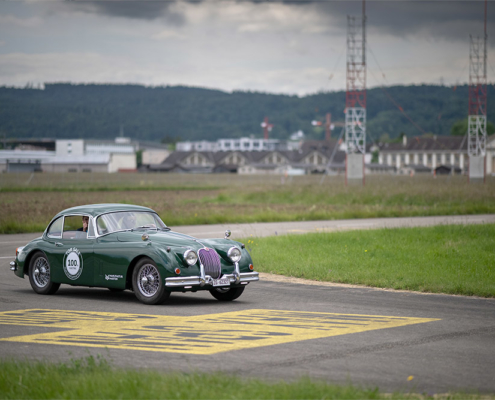 Jaguar auf Flugfeld Ein grüner Jaguar XK 150 fährt über eine Flughafen-Fahrbahn. Gelbe Marierungen auf der Fahrbahn, im Hintergrund Baumarkierungen. Auf der Seite des Jaguars ist eine Beschriftung angebracht, die darauf hinweist, dass es sich um ein Elektroauto handelt.