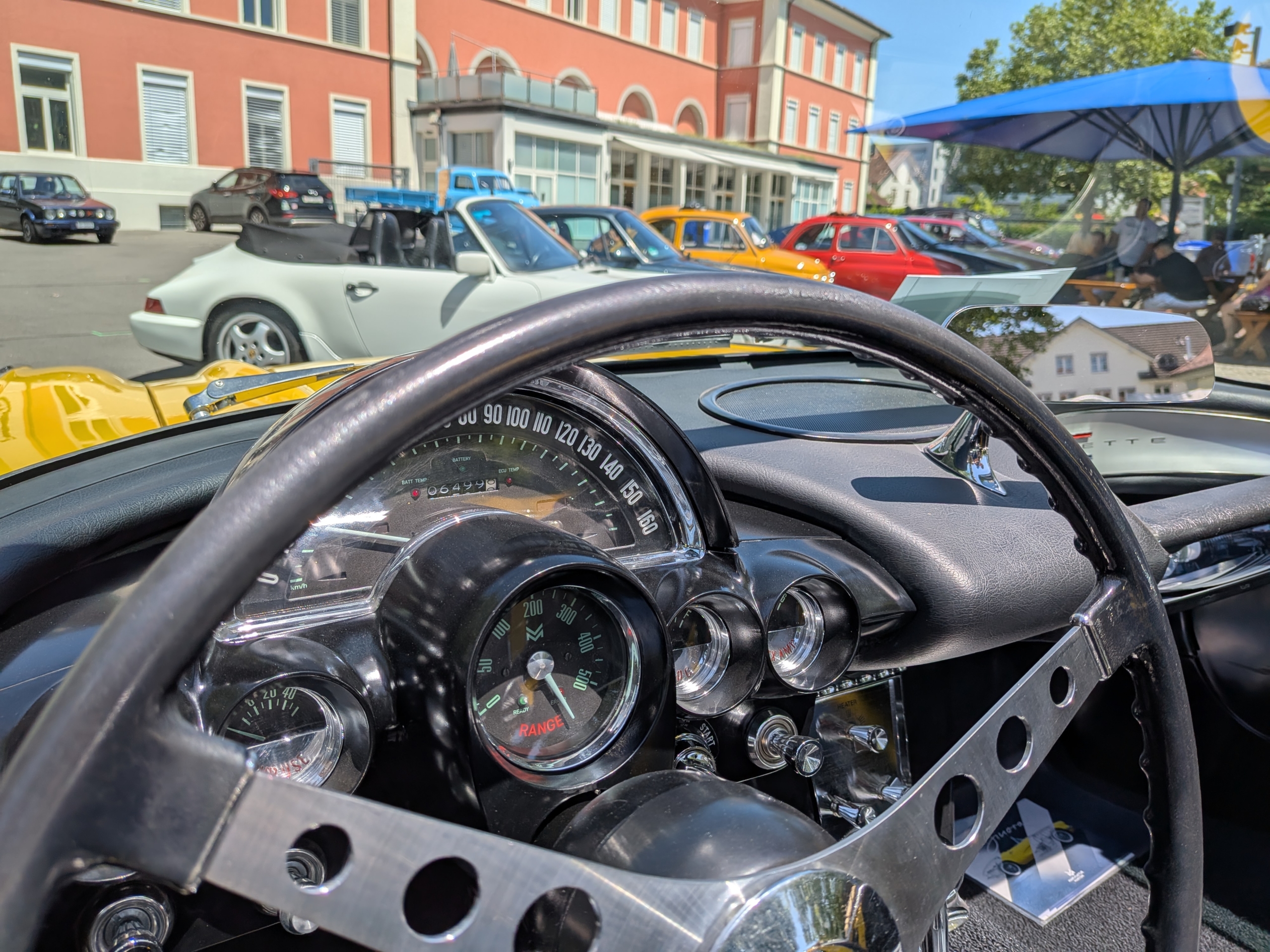 WheelJam mit Dinora Blick über das Lenkrad einer Corvette C1. Die Armaturen verraten, dass es sich hier um ein Fahrzeug handelt, dass zum Elektroauto umgebaut wurde. Denn an Stelle des Tourenzählers zeigt die klassische Rundanzeige die Restreichweite an. Im Hintergrund sieht man diverse Fahrzeuge. Rechts eine Reihe mit diversen Fahrzeugen. Zwei Porsche 911 in weiss und grau, zwei Fiat Nuova 500 in gelb und rot sind gut erkennbar, dahinter lassen sich zwei Alfa Romeo GTV in rot und blau sowie eine Charleston-Ente erahnen.