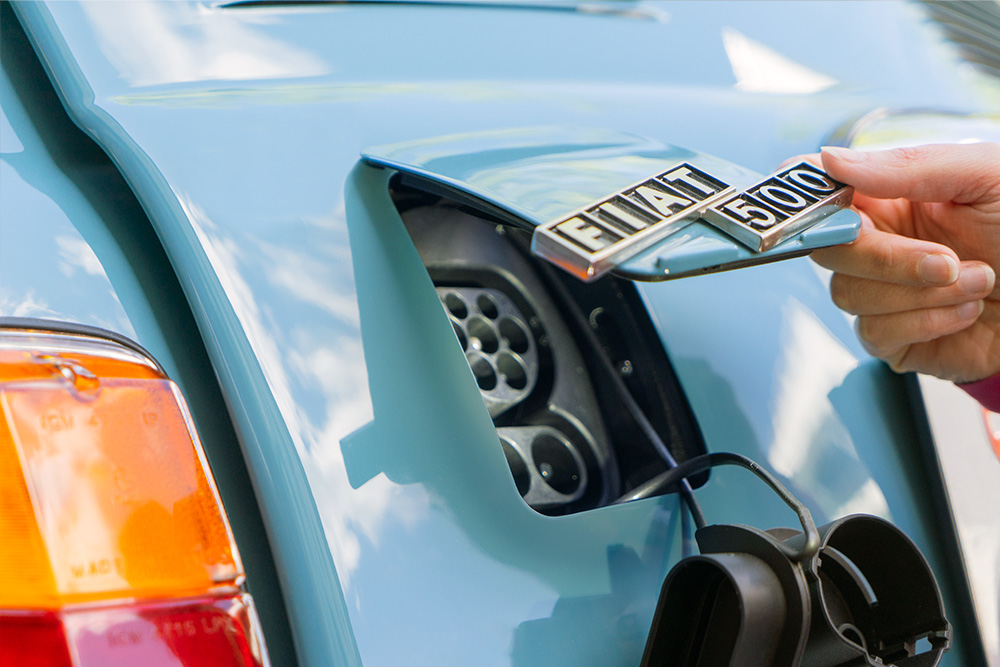 The finished charging flap Eine Öffnung am Heck des blau lackierten Fiat 500, die mit einer Klappe in Wagenfarbe verschlossen werden kann. Im Bild ist diese halb geöffnet, an ihr ist das Logo "Fiat 500" angebracht.