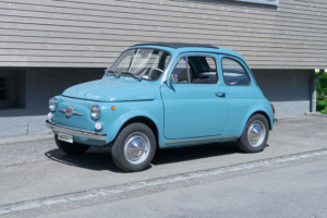 Ein mittelblauer Fiat Nuova 500 steht vor einem Gebäude mit Holzfassade. Das Fahrzeug ist von der Seite fotografiert.
