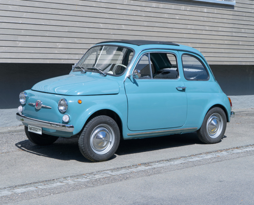 Fiat 500, newly restored and electrified Ein mittelblauer Fiat Nuova 500 steht vor einem Gebäude mit Holzfassade. Das Fahrzeug ist von der Seite fotografiert.